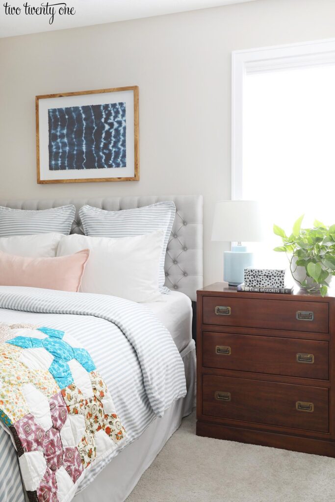 1986 Ethan Allen campaign-style night stand, bed with gray tufted headboard, pillows, blue and white striped duvet and vintage quilt. Shibori wall art above bed.